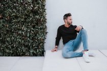 Adulto bello elegante uomo pensoso in abbigliamento casual seduto sul pavimento e guardando lontano vicino alla parete grigia — Foto stock