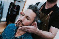 Barbeiro fazendo massagem facial para bonito macho elegante com olhos fechados sentado na cadeira — Fotografia de Stock