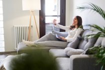 Giovane donna allegra che utilizza il computer portatile e prende selfie con il telefono cellulare sul divano a casa — Foto stock