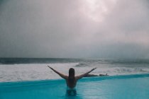 Vista trasera de mujer joven con las manos levantadas descansando en el agua de la piscina en la costa del mar tormentoso - foto de stock