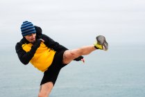 Hombre barbudo adulto en ropa deportiva practicando entrenamiento de kickboxing en la costa del mar - foto de stock