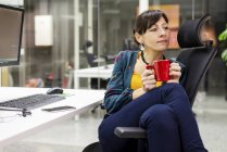 Pensativo gerente femenino con taza de bebida caliente descansando en la silla en la oficina - foto de stock