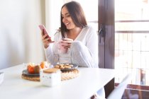 Mujer feliz joven usando el teléfono móvil y desayunando en la mesa cerca de la ventana en casa - foto de stock