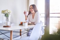 Sorrindo jovem mulher tomando café da manhã na mesa perto da janela em casa — Fotografia de Stock