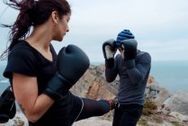 Uomo e donna in guantoni da boxe che si picchiano a vicenda mentre stanno in piedi sulla scogliera contro mare e cielo — Foto stock