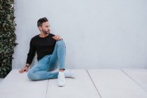 Adult handsome elegant pensive male in casual wear sitting on floor and looking away near grey wall — Stock Photo