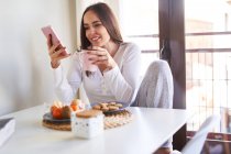 Junge glückliche Frau benutzt Handy und frühstückt zu Hause am Tisch neben dem Fenster — Stockfoto
