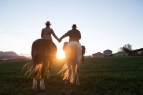 Rückansicht von Mann und Frau auf Pferden und Händchen haltend gegen den Sonnenuntergang auf der Ranch — Stockfoto