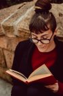 Молодая элегантная женщина в очках опирается на каменную стену и читает книгу — стоковое фото