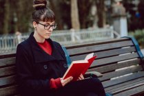 Young elegant woman in eyeglasses reading book while sitting on bench in city park — Stock Photo