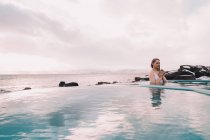 Giovane donna con gli occhi chiusi meditando in acqua di piscina vicino rocce e cielo nuvoloso — Foto stock