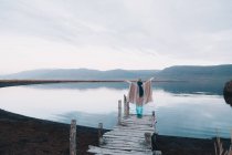 Back view of young woman in poncho with upped hands standing on dock near amazing water surface between hills — Stock Photo