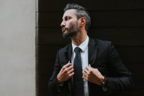 Adulto bell'uomo d'affari elegante in abito formale di regolazione giacca e guardando lontano vicino alla parete grigia — Foto stock