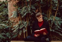 Jeune femme élégante dans les lunettes de lecture livre et assis sur le banc dans le jardin de la ville — Photo de stock