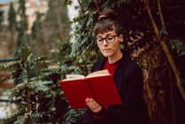 Young attractive elegant woman in eyeglasses reading book in city garden — Stock Photo