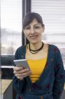 Porträt einer Managerin, die ihr Smartphone am Fenster hält, während sie im modernen Büro arbeitet — Stockfoto
