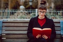 Young attractive elegant woman in eyeglasses reading book and sitting on bench in city garden — Stock Photo