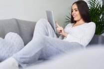 Молода щаслива жінка використовує цифровий планшет і відпочиває на дивані вдома — стокове фото