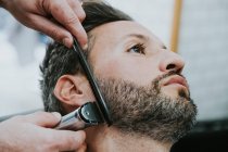 Primer plano del peluquero con peine y recortadora cortando la barba del macho sentado en la peluquería sobre fondo borroso - foto de stock