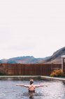 Back view of young woman resting in water of pool against wooden fence in nature with mountain on background — Stock Photo