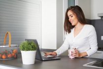 Smiling young woman with cup of coffee using laptop at kitchen counter at home — Stock Photo