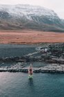 Visão traseira da mulher com as mãos levantadas nadando na água perto de rochas na costa e montanha na neve entre terras selvagens — Fotografia de Stock