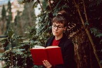 Joven atractiva mujer elegante en anteojos libro de lectura en el jardín de la ciudad - foto de stock