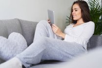 Молода щаслива жінка використовує цифровий планшет і відпочиває на дивані вдома — стокове фото