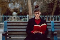 Молода приваблива елегантна жінка в окулярах читає книгу і сидить на лавці в міському саду — стокове фото