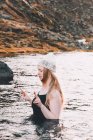 Junge blonde Frau in Hut und Badeanzug mit geschlossenen Augen meditiert in der Wasseroberfläche in der Nähe der felsigen Küste — Stockfoto