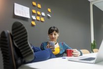Manager donna con tazza di bevanda calda utilizzando lo smartphone mantenendo le gambe sulla scrivania in ufficio — Foto stock