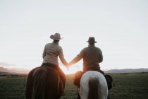 Вид ззаду чоловік і жінка верхи на конях і тримаються за руки проти заходу сонця небо на ранчо — стокове фото