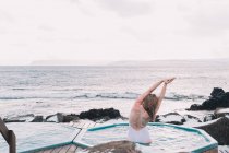 Visão traseira da mulher loira com as mãos levantadas relaxando na água da piscina perto de rochas em nublado — Fotografia de Stock
