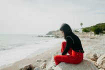 Стильна жінка, дивлячись вниз, сидячи на скелі на пляжі — стокове фото