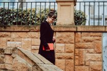 Молодая элегантная женщина в очках держит книгу и спускается по лестнице перед зданием в городе — стоковое фото