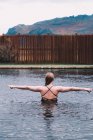 Visão traseira da jovem que descansa na água da piscina contra cerca de madeira na natureza com montanha no fundo — Fotografia de Stock
