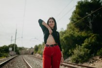 Pretty young woman in stylish outfit lifting sweater to show mountains tattoo while standing on blurred background of rails — Stock Photo