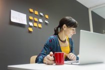 Бізнес-леді з чашкою гарячого напою переглядаючи ноутбук під час роботи в сучасному офісі — стокове фото