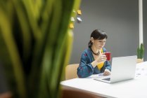 Mujer de negocios con taza de bebida caliente portátil de navegación mientras trabaja en la oficina moderna - foto de stock