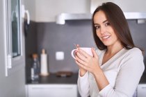 Retrato de una joven mujer sonriente sosteniendo taza en la cocina moderna en casa - foto de stock