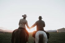 Back view of man and woman riding horses and holding hands against sunset sky on ranch — Stock Photo