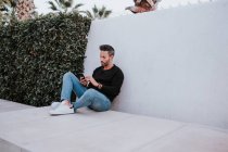 Гарний елегантний щасливий чоловік у повсякденному одязі, використовуючи мобільний телефон і сидячи біля сірої стіни — стокове фото
