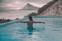 Вид ззаду молода жінка з простягнутими руками, що стоять у воді басейну біля скель на узбережжі і штормового моря — стокове фото