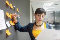 Businesswoman holding document and writing on sticky notes while standing near gray wall in office — Stock Photo