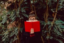 Jeune femme élégante avec des lunettes regardant la caméra tout en couvrant le visage avec un livre dans le jardin de la ville — Photo de stock