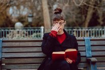 Giovane donna elegante in occhiali libro di lettura mentre seduto sulla panchina nel parco della città — Foto stock