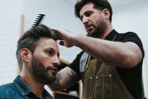 Da sotto barbiere pettinando capelli di bel maschio elegante seduto in barbiere — Foto stock
