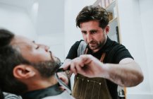 Barber with comb and trimmer cutting beard of male sitting in barbershop on blurred background — Stock Photo
