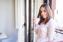 Lächelnde junge Frau mit Tasse am Fenster zu Hause und Blick in die Kamera — Stockfoto