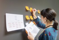 Geschäftsfrau hält Dokument in der Hand und schreibt auf klebrige Zettel, während sie in der Nähe einer grauen Wand im Büro steht — Stockfoto